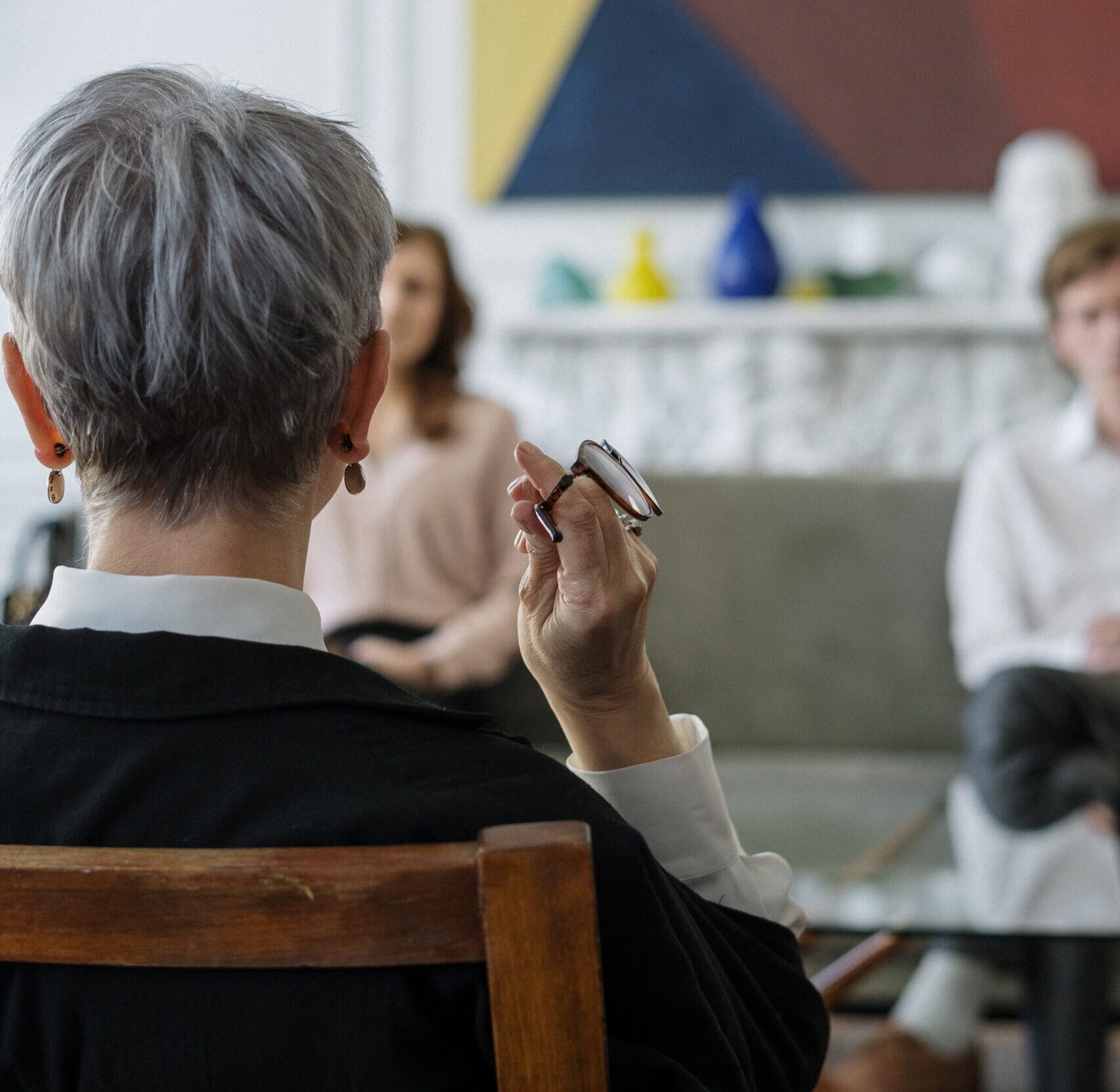 Woman sitting in a chair holding a pair of glasses and speaking with two other people