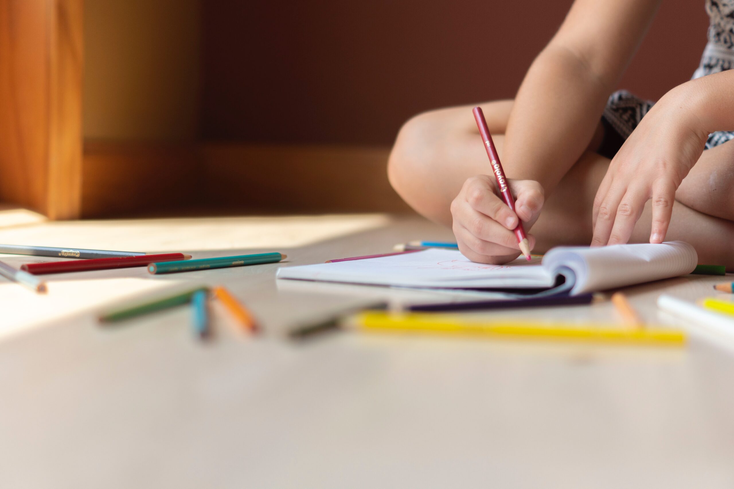 Young child's hands writing in a book on the floor surrounded by colouring pencils